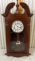 Wooden wall clock with decorative carved crown and columns, including pendulum visible behind glass panel.