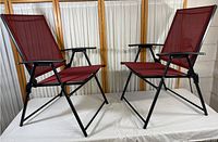 Two folding outdoor chairs with black metal frames and maroon mesh fabric seating and backs, shown together on a white surface with a wooden screen background.