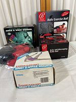 Set of ankle and wrist weights, Bally exercise ball, and self-massaging roller ball in original packaging on table.