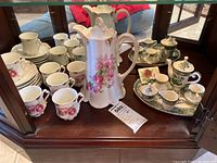 Photo showing full grouping: chocolate pot with pink floral design, six matching cups and saucers, green and white demitasse tea set on tray.
