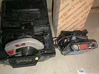 Craftsman circular saw and belt sander shown in black carrying case on a concrete floor with box and other objects in background.