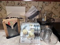 Food processor base with clear plastic bowl, cord, a clear disc holder with multiple discs, a manual bagel slicer with wooden handles, boxed disc set, instruction manual.