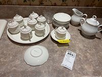Full lot displayed showing eight lidded cups on tray with saucers, plus Dansk sugar bowl, cream pitcher, and ribbed clear glass pitcher.