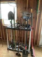 Full view of garden tools and plastic holder showing shovels, pick, broom, and rakes in used condition.