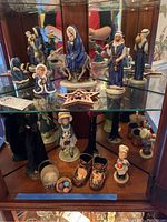 Display cabinet with Goebel nativity figures, Holly Hobbie figurines, bronzed baby shoes, tall ladies figurines, and metal basket arranged on glass shelves.