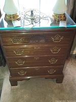 Front view showing cherry wood nightstand with four drawers, brass hardware, custom beveled glass top protector and lamps on top