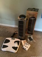 Lasko ceramic tower heater standing in front of its original box with remote control in plastic bag. Digital scale with black and white design placed beside heater.