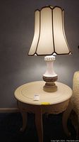 Full view of ceramic base table lamp with beige fabric shade standing on round wood table.