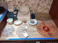 Photo of various porcelain, glass, and decorative items including Lenox bowls, colored glass plates, a blue glass pitcher, and shell napkin rings arranged on a surface.
