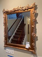 Full view of the rectangular gold tone framed mirror hung on wall near staircase showing size and overall design with leaf decorations.