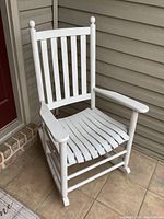Full view of white wood rocking chair on tile porch next to brick and wall, showing vertical slat backrest and curved rocker base.