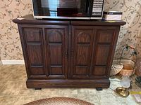 Closed wooden server with paneled doors, dark wood finish, placed on tiled floor with microwave on top.