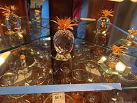 Three faceted Swarovski crystal pineapples with gold-tone tops on glass shelf with mirrored surfaces beneath.