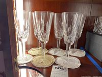 Six clear Waterford crystal champagne glasses displayed on a wooden shelf with lace coasters, showing their faceted cut pattern and stems.