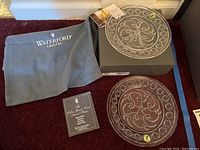 Two matching Waterford Society Celtic Spiral Knot accent plates, one partially resting on box, one on carpet, with Waterford Crystal branded carrying bag and pamphlet included