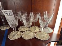 Six matching Waterford stemware glasses with lace coasters on wooden shelf.