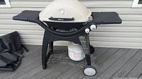 Weber Q Series grill with cream lid, black side trays, and propane tank underneath. Grill cover folded on side.