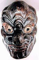 Front view of the antique Japanese paper mache theater mask showing the carved and hand painted details of a fierce face in dark colors with visible wear.