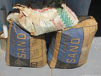 Two 60 lb all-purpose sand bags stacked side by side with a partially crumpled bag of ammonium sulfate on top, placed on tiled flooring near a blue wall.