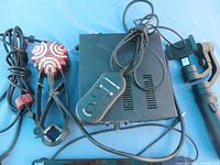 Overall view of the amplifier unit with attached cables, remote control, mounting clamp, and vibrating transducer device on a blue surface.