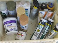 Photo showing assortment of chemical products including DAP concrete sealant tubes, SimpleGrout pre-mixed grout, Rust-Oleum spray paints, and a spray cleaner bottle inside a white plastic basket.