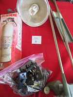 Lot displayed on red surface showing Kidde fire extinguisher in box, clamp light with bulb, vintage wooden drafting tool, and bag of light bulb holders.