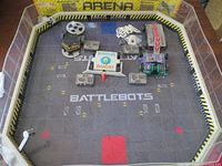 Top-down view of Hex Bug Battlebots arena with 4 Battlebots and controllers inside. Various robot designs and arena floor logo visible.