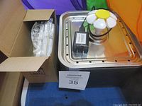 Stainless steel kitty fountain with attached flower-shaped water sprayer on top, along with a black power adapter and some boxed accessories.