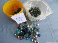Collection of assorted glass marbles in yellow cup, plastic bag, and scattered on surface