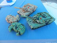 Two remote control tanks with camouflage and their matching controllers on a blue surface.