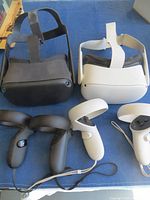 Photo showing two Mediquest VR headsets, black v1 model and white v2 model, each with their respective two controllers, placed on blue cloth surface.