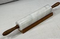 White marble rolling pin with wooden handles lying on a wooden base holder on a plain surface.