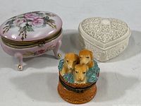 Three small decorative lidded trinket boxes: pink porcelain floral box on legs, white heart-shaped ceramic box, and dog head ceramic box.