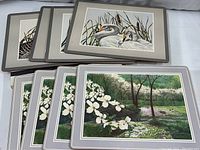 Photo showing eight solid wood placemats arranged with four placemats featuring a waterfowl scene including swans and four placemats featuring a nature scene with flowering dogwood trees.