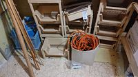 White plastic bin containing an orange extension cord, placed on the floor in front of stacked beige baskets and wooden frame pieces.