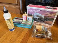Photo shows BeDazzler Rhinestone & Stud Setter boxed kit alongside a bottle of paper adhesive, several bars of soap in a small basket, and a clear plastic bag with assorted perfume bottles and small items.