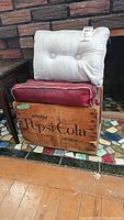 Vintage wooden Pepsi-Cola crate with visible branding, holding five cushions; two cushions are specific, one gray with buttons and one red, both stained.