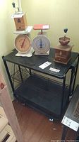Black metal utility cart with two wire shelves and solid top, situated near a brick wall and wooden floor, with vintage kitchen scales and a manual coffee grinder placed on top (not included).