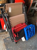 Set of folding chairs including a beige fabric lounge chair with black frame, a red folding seat with a black pouch and red strap, and a small blue plastic folding seat.
