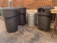 Front and side view of the three Rubbermaid garbage bins and one aluminum garbage can lined up outside near a brick wall.
