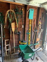 Photo showing various garden tools hung on a brown wooden wall including brooms, rake, gardening forks, shovels, weed pullers, a small wooden ladder, wire fencing, and a yellow level.