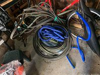 Photo showing three garden hoses coiled on the ground near a red bicycle. One hose is tan with green stripes, one is dark green, and one is blue expandable hose. The blue sprinkler with a metal base is visible to the right.
