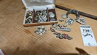 Box containing multiple assorted brooches and earrings spread out on a wooden surface, with varying designs including floral and hand shapes.