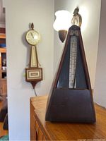 Wooden metronome placed in front of MCM Seth Thomas electric wall clock on wall, showing overall shapes and condition.
