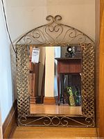 Full view of the metal framed wall mirror positioned on the floor, showing the woven metal design on sides and decorative scrollwork on top and bottom of the frame.
