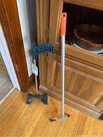 Two reacher grabber tools positioned beside a wooden cabinet on a hardwood floor, one with a blue handle and claw, the other with an orange handle and claw.