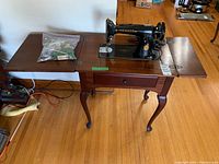 Full view of sewing machine mounted in wooden table with both drop leaves open and accessory bag on top.