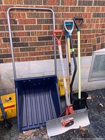 Wide view of the snow removal tools including shovels, ice chippers, and broom against a brick wall background.