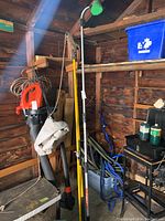 View of the trimmer, brush hose head, and other tools in a wooden shed corner.