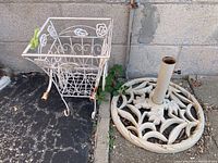 Photo showing both items: the metal planter stand and the metal umbrella base placed on the ground against a wall.